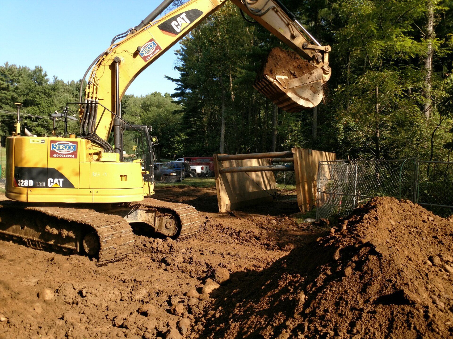 Yellow excavator digging in dirt near a fence, trees in background on a sunny day.