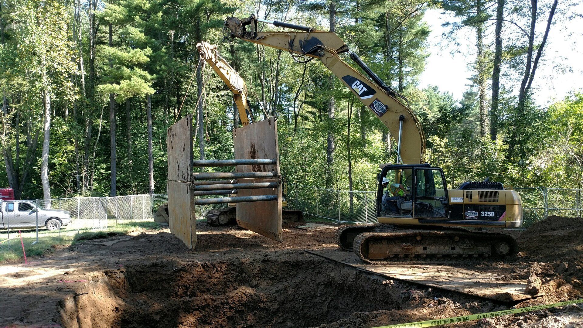 Excavator with trench box in dirt pit, with trees and a truck in the background.