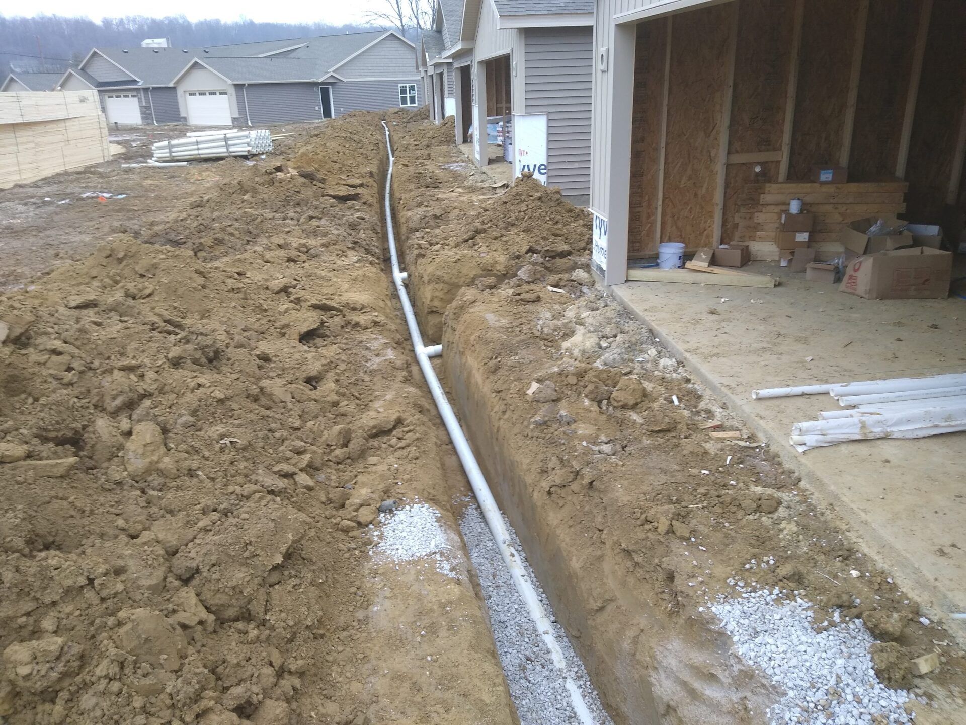 Trench with white drainage pipe running alongside a building under construction, surrounded by dirt and gravel.