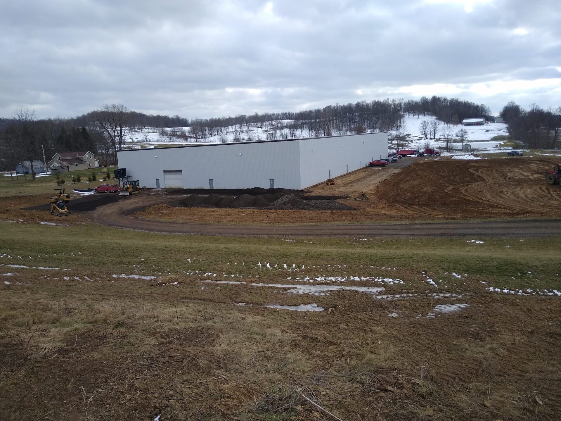 A large, white industrial building sits near a snow-covered hill on a cloudy day.