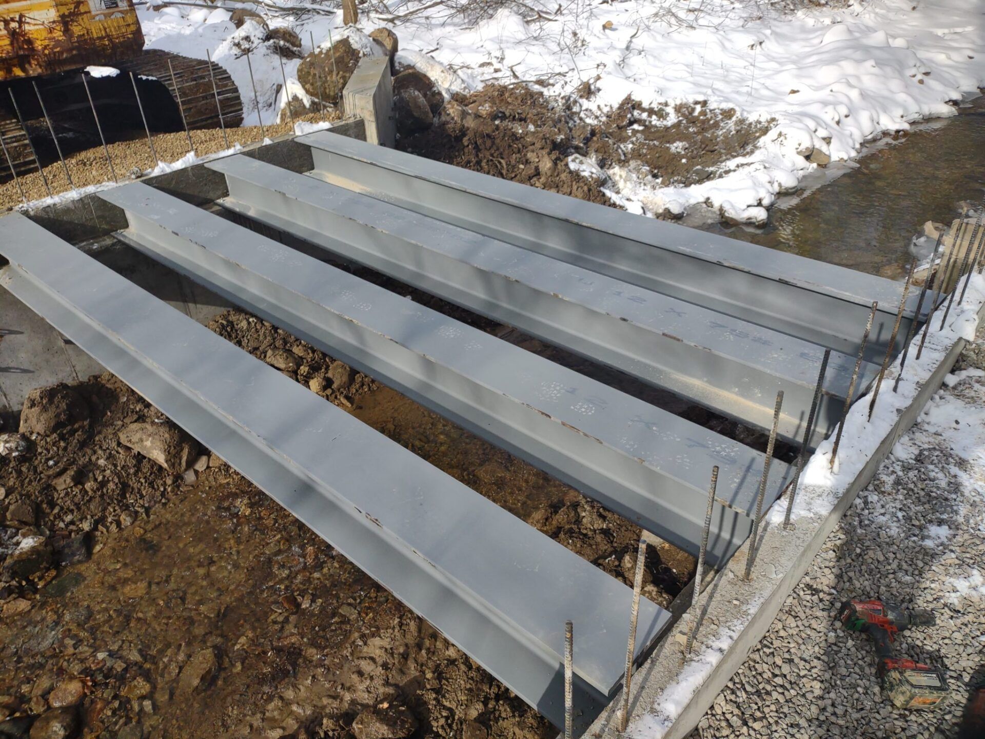 Construction site: Steel beams in a concrete frame, prepared for installation, with an excavator in background.