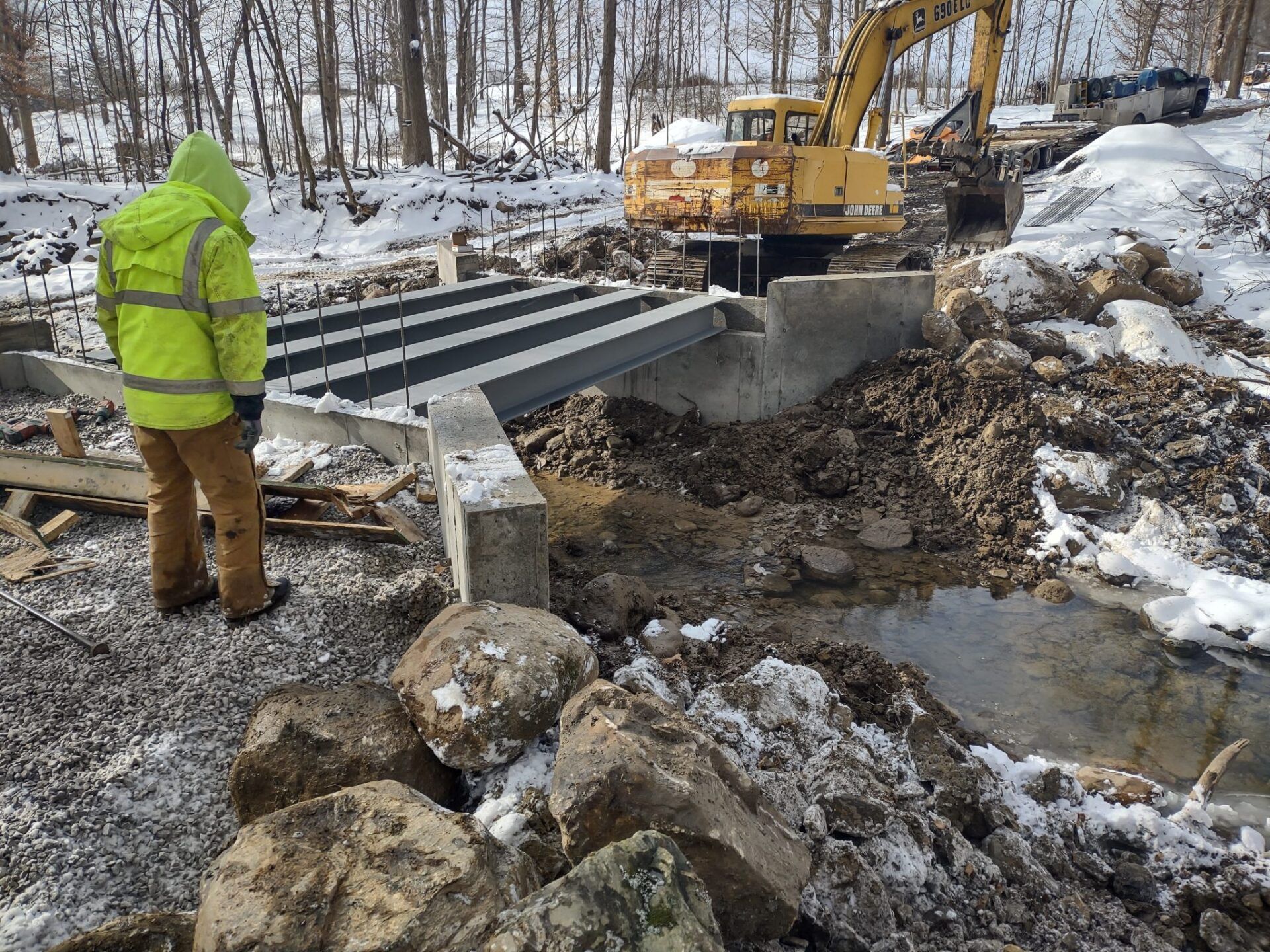 Construction site: worker in yellow vest observes bridge construction in snowy environment with excavator.