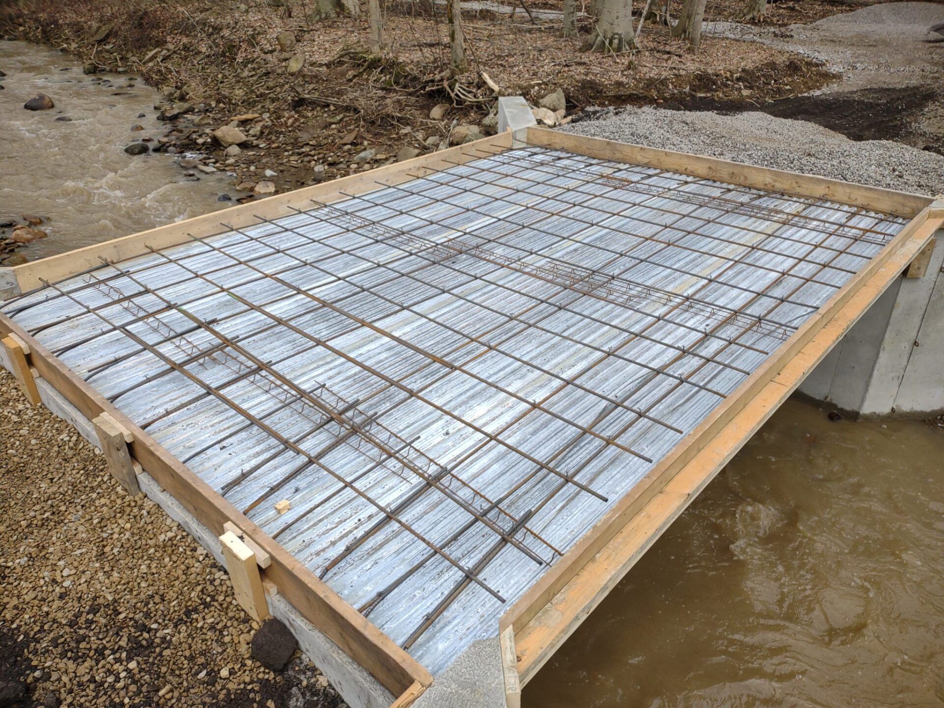 Bridge construction: Wooden formwork with rebar grid over a stream.