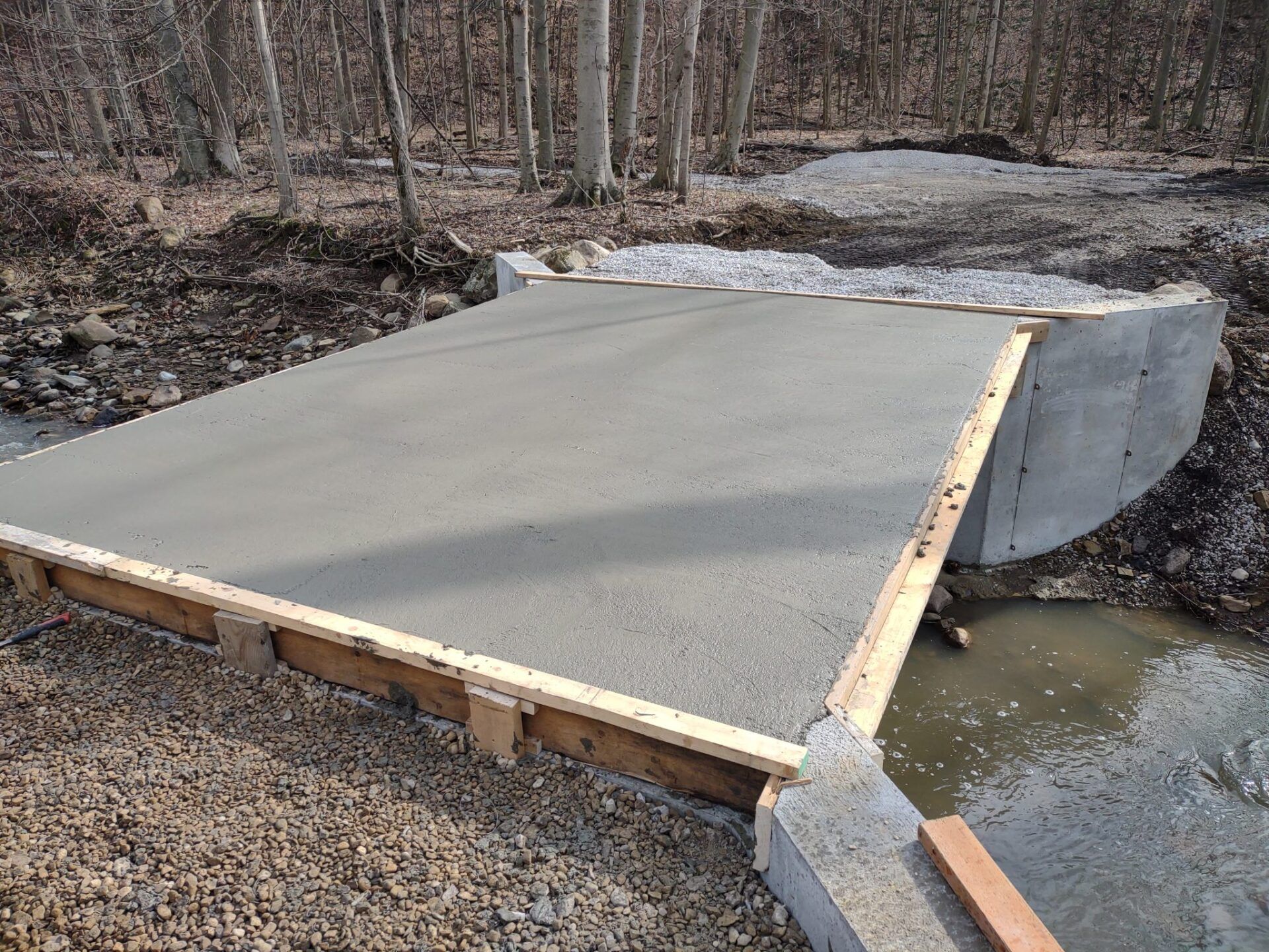 Newly poured concrete bridge over a small stream, surrounded by gravel and trees.