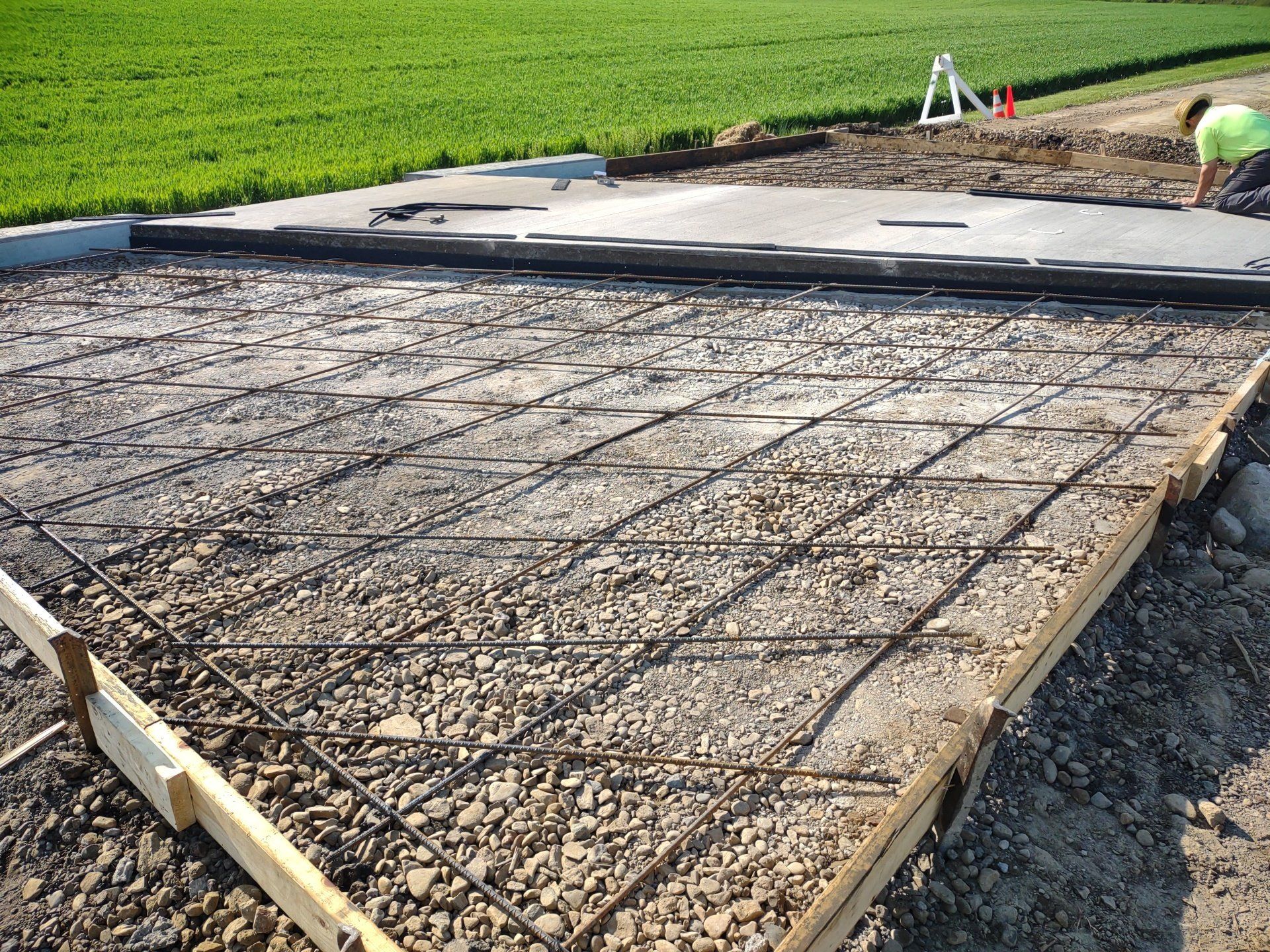 Concrete slab construction, rebar grid visible; worker in background near green field.