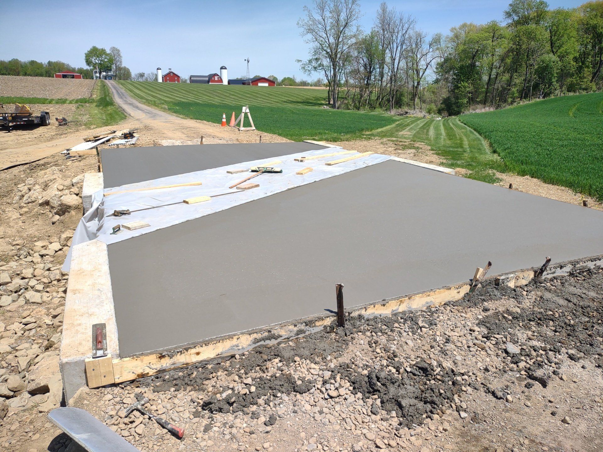 Freshly poured concrete slab with wooden formwork on a construction site, surrounded by fields and a distant road.