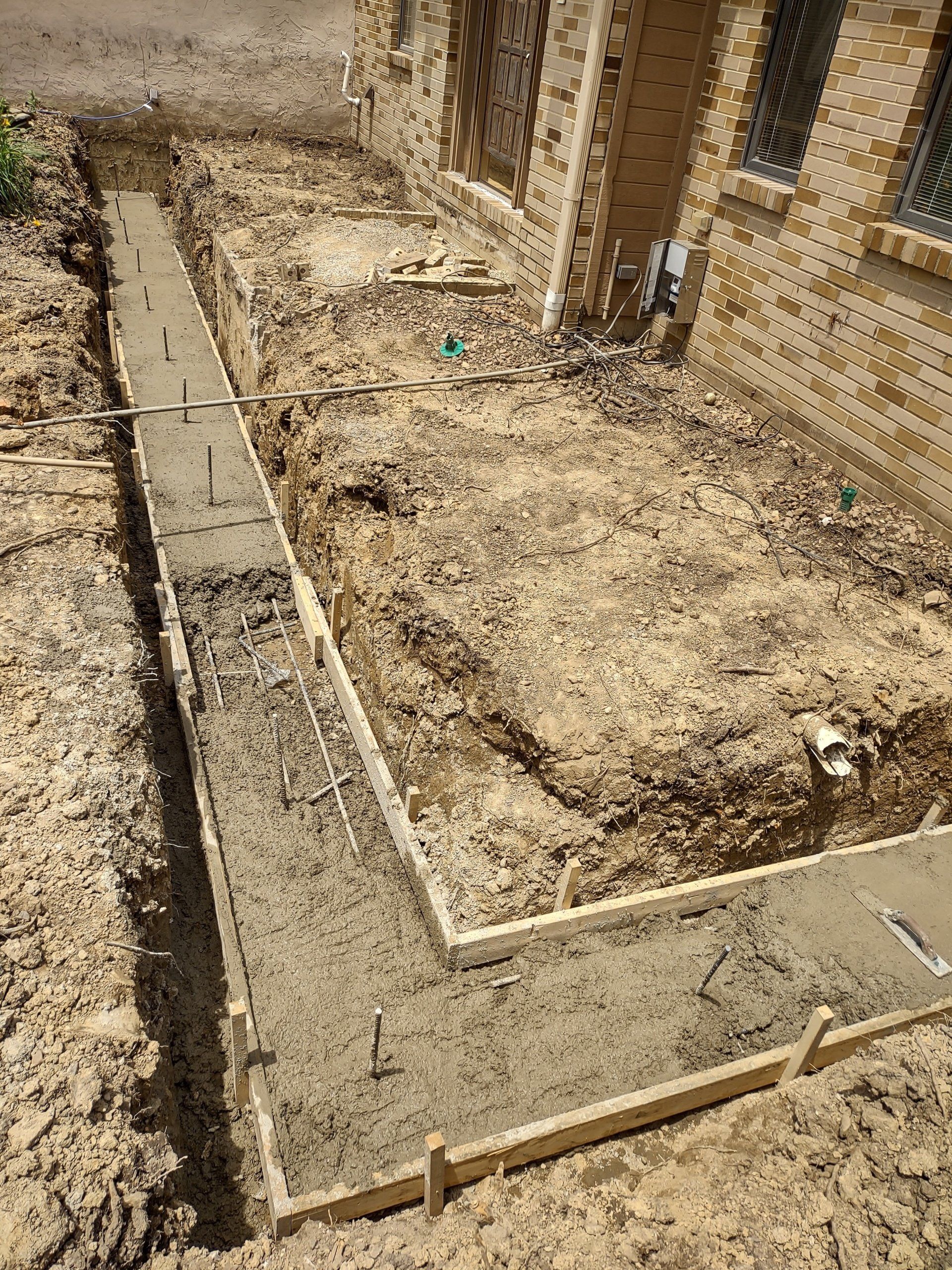 Freshly poured concrete foundation along a house. Wooden forms and rebar are visible.