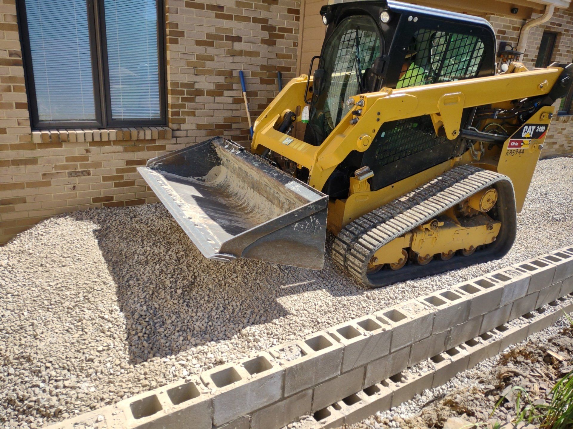 Yellow skid steer with bucket on gravel, near a brick building and a cinder block wall.