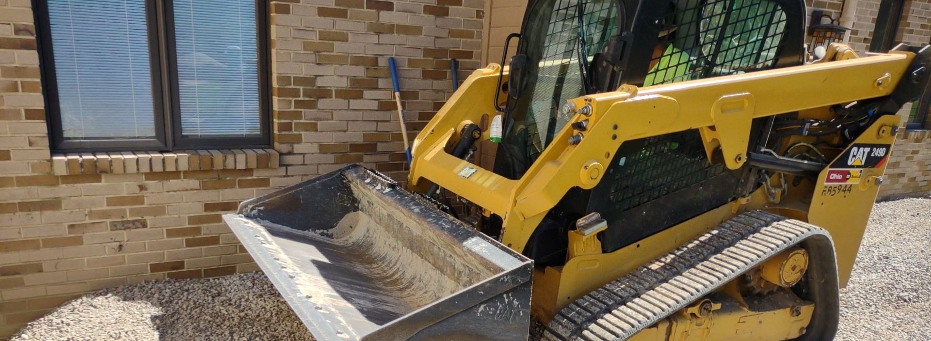 A yellow skid steer loader with a full bucket is near a brick building.