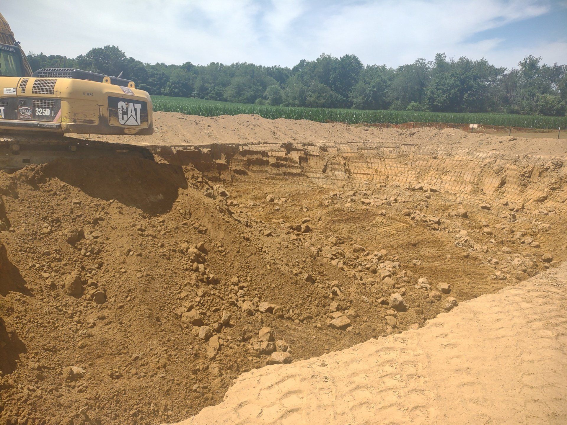 Yellow excavator digging in a large dirt pit; trees in background under a blue sky.