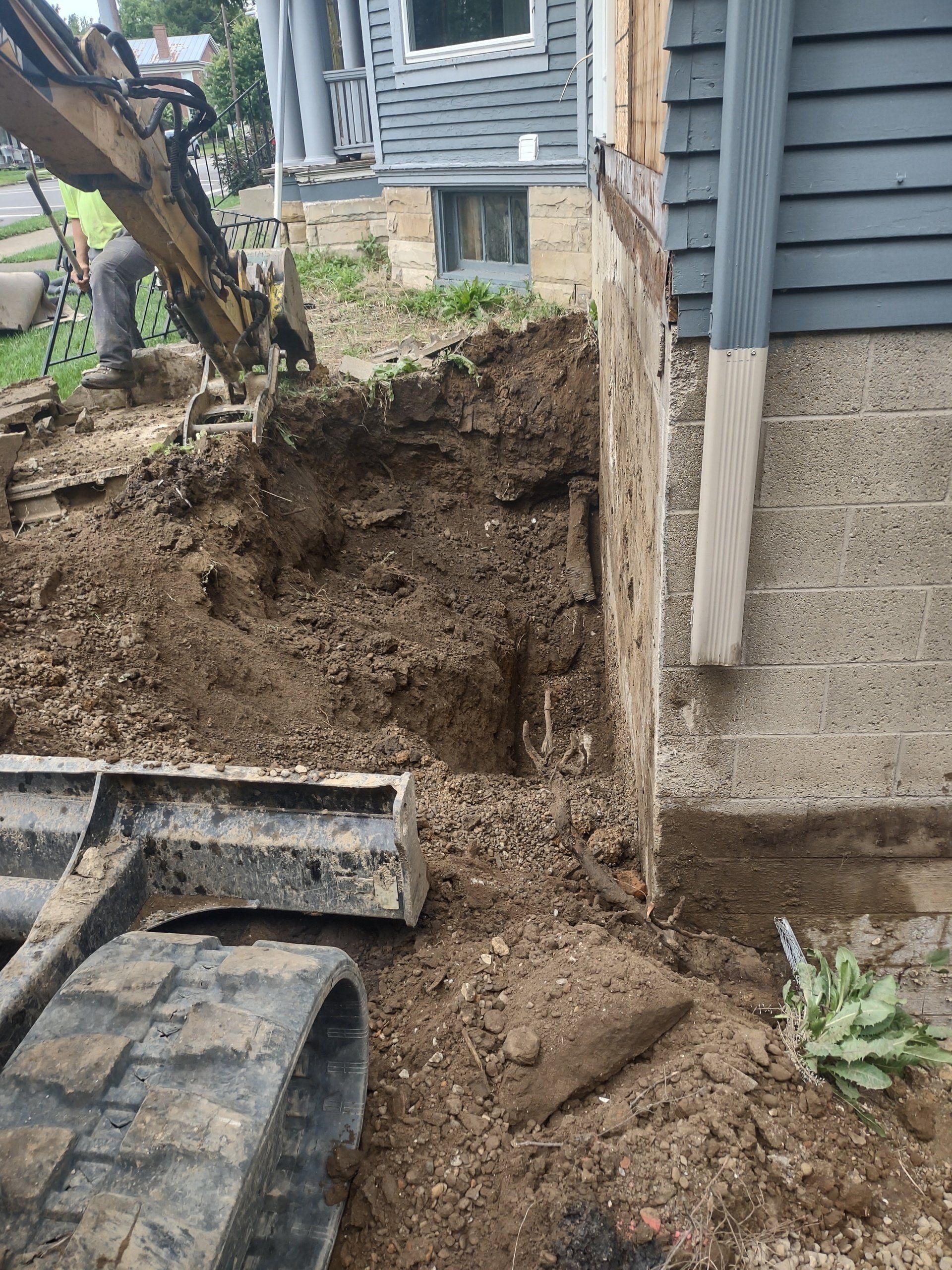 Excavator digging near a house foundation. Soil removed, exposing concrete wall. Cloudy day.