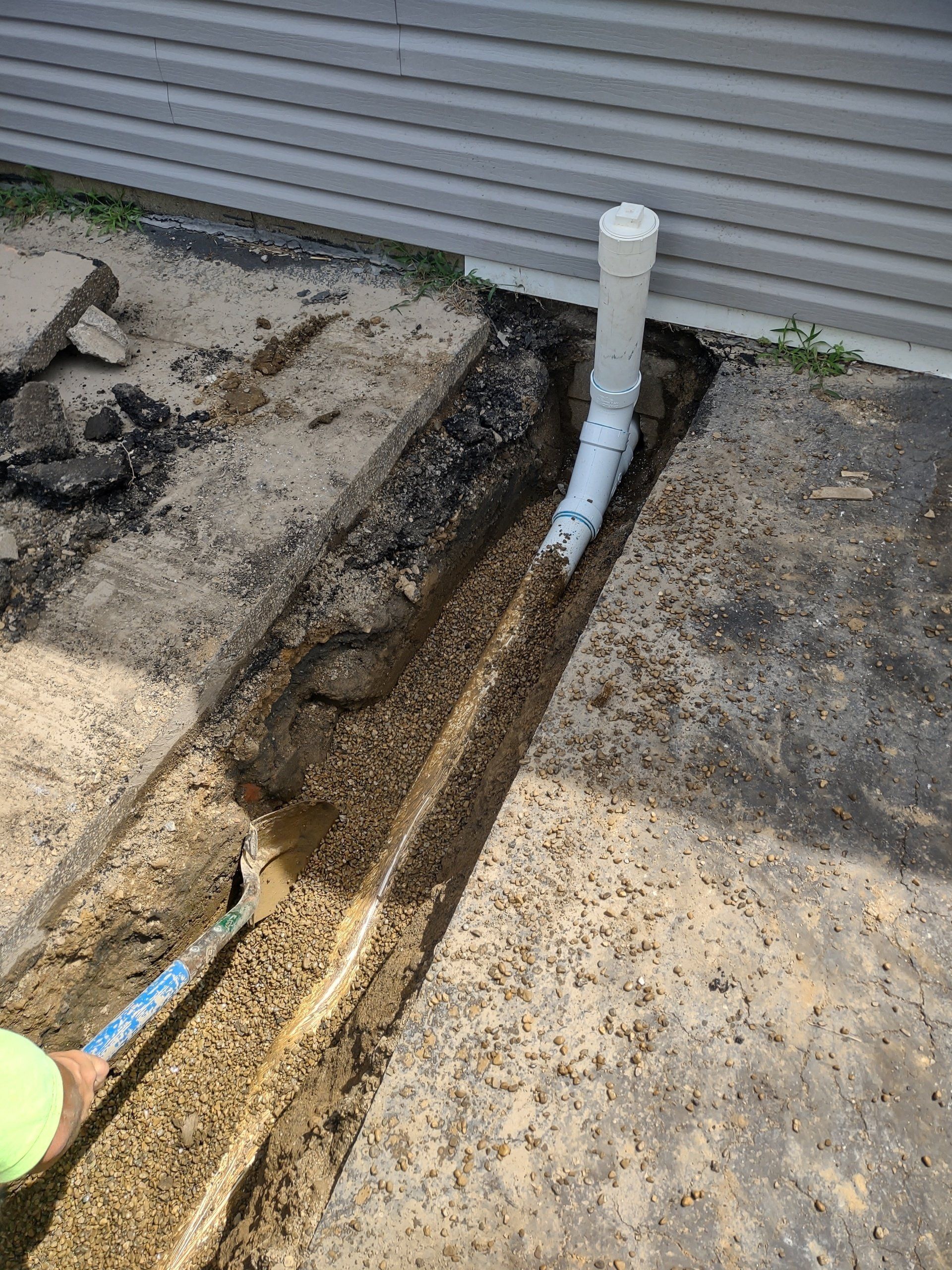 Trench dug near a building, with a white PVC pipe extending into it; a person with a shovel is visible.