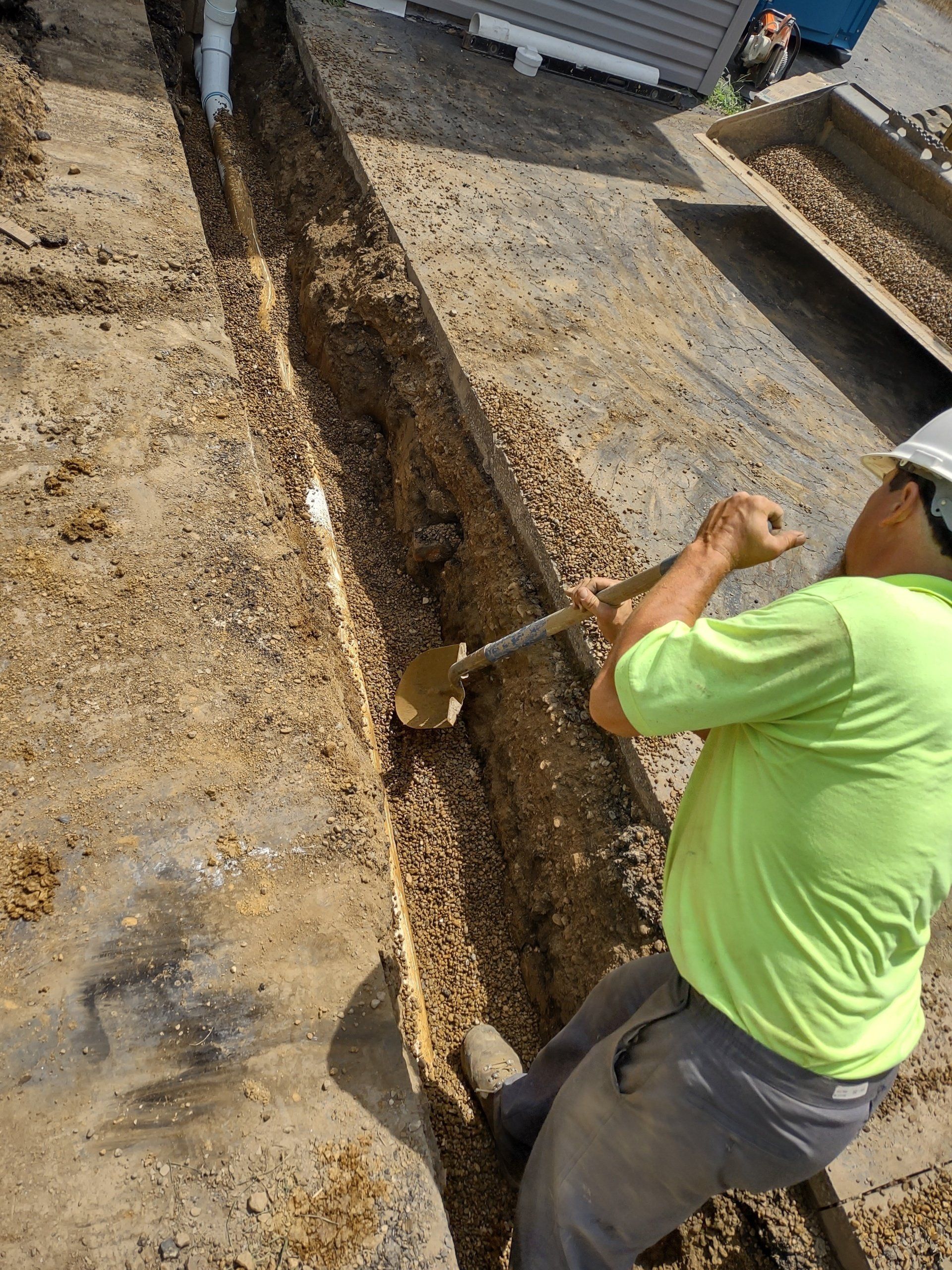 Construction worker digging in a trench with a shovel, surrounded by gravel.