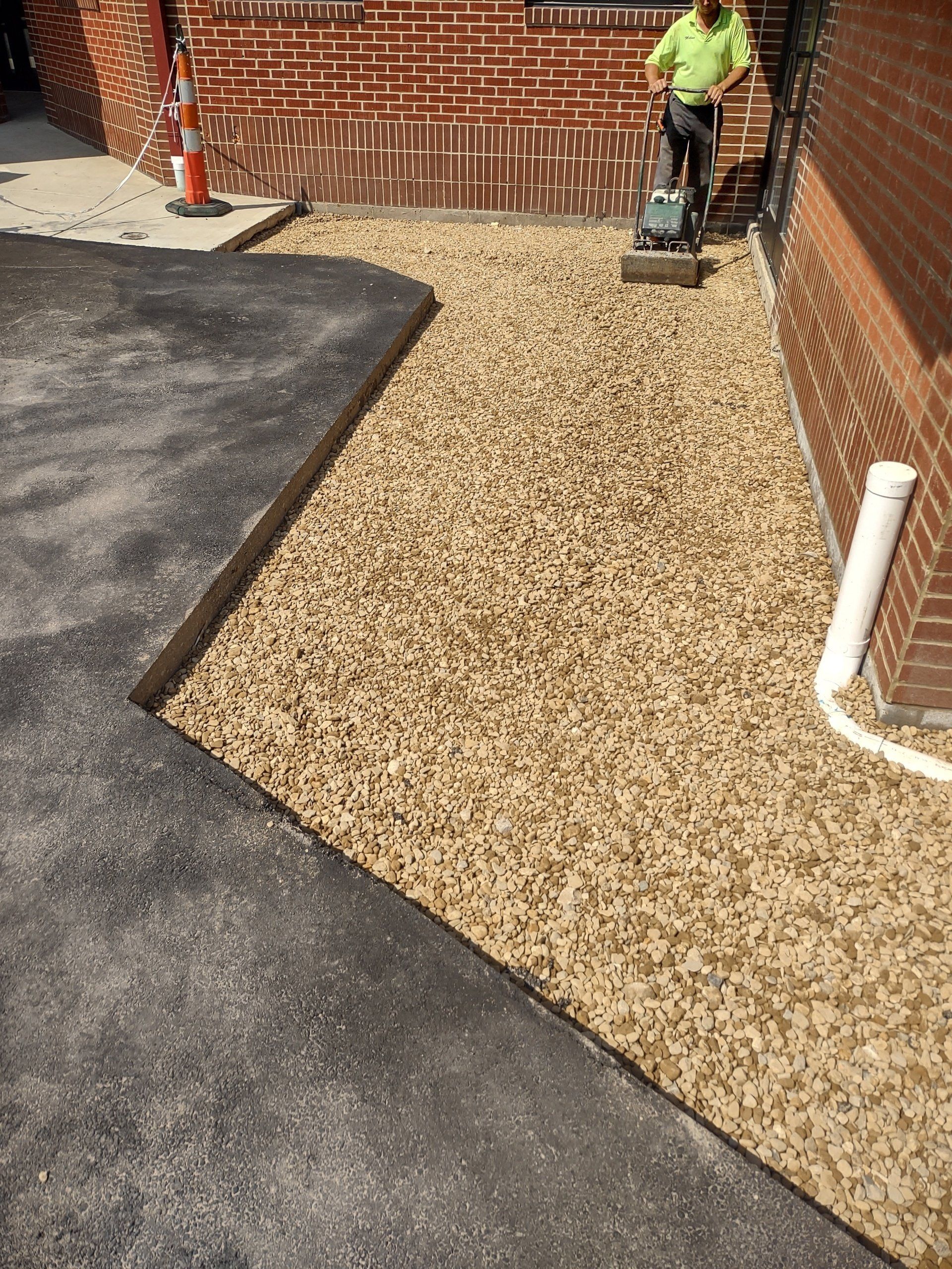 A person using a machine to smooth gravel; next to asphalt walkway and brick building.