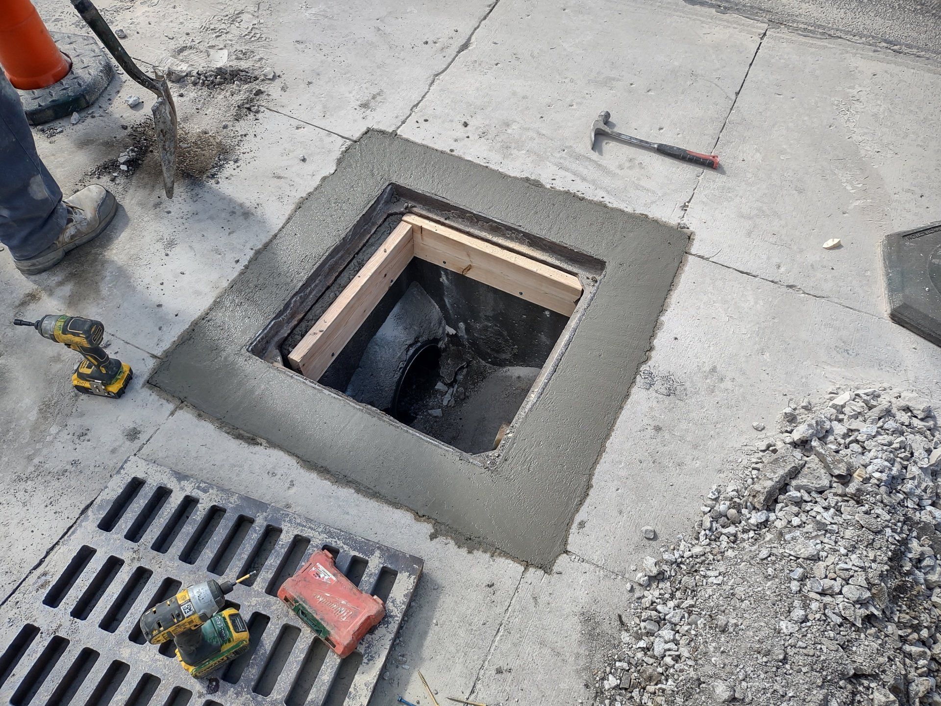Construction site: Open utility access in concrete, surrounded by fresh cement. Drain grate and tools are present.