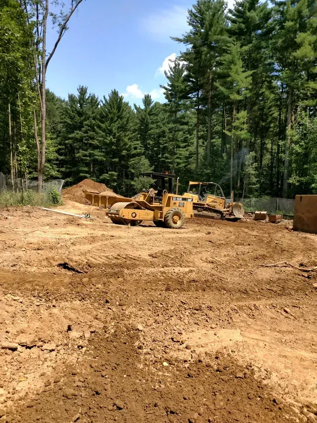 Construction site with heavy machinery on a dirt lot, surrounded by trees under a bright sky.