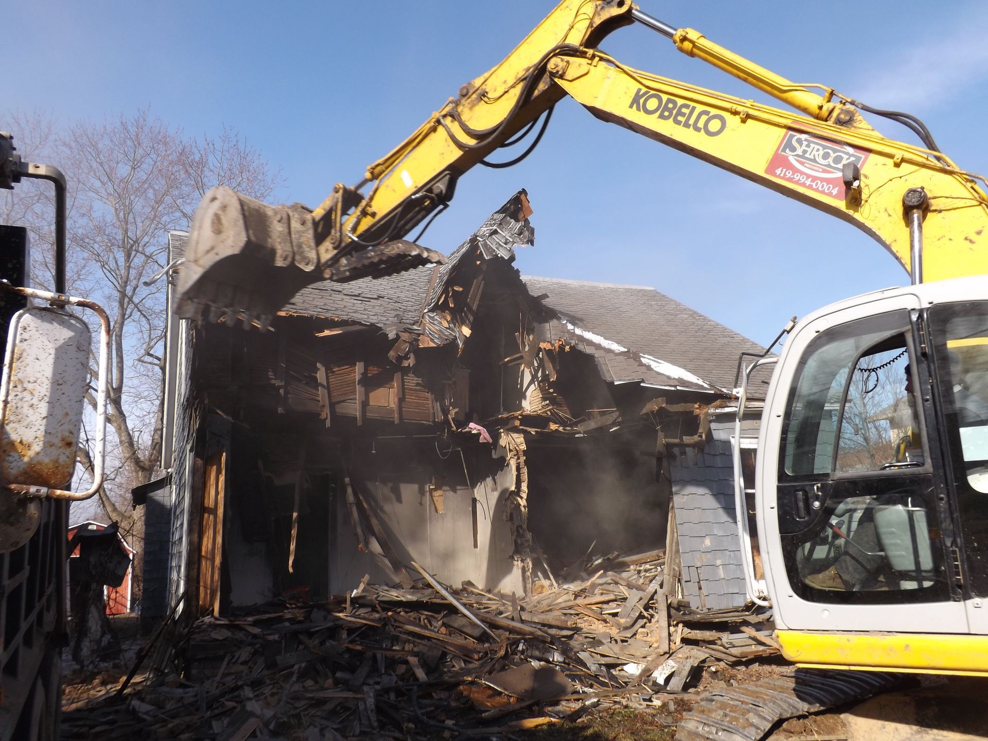 Yellow excavator demolishing a building; dust and debris visible.