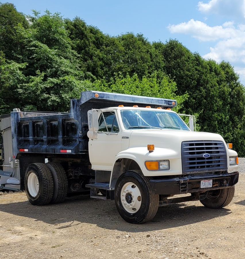 White Ford dump truck parked on gravel, with trees in the background under a blue sky.