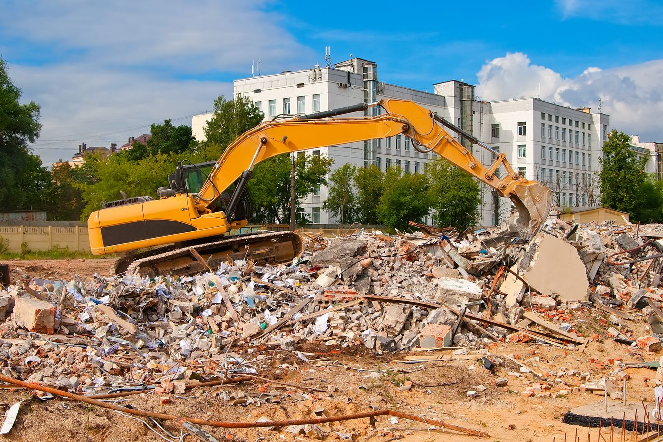 Yellow excavator demolishing a building, surrounded by rubble under a blue sky.
