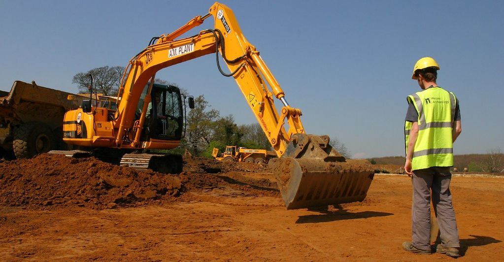Yellow excavator on dirt, worker in safety vest and helmet observes on construction site under blue sky.