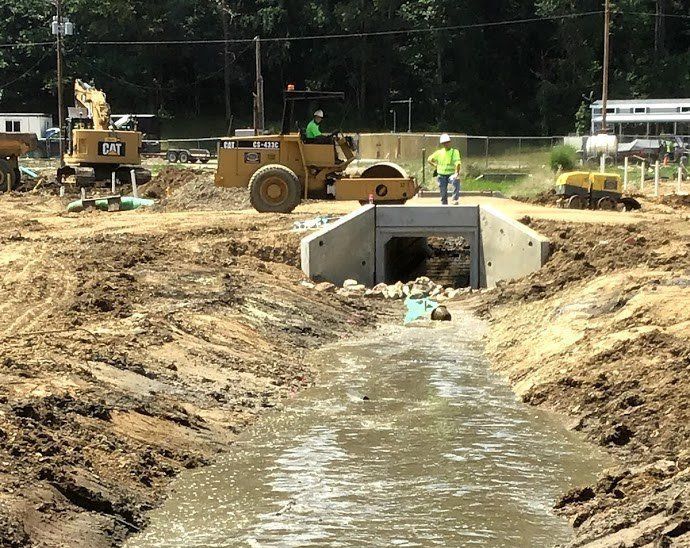 Construction site with culvert, heavy machinery, and workers. Muddy terrain, overcast day.