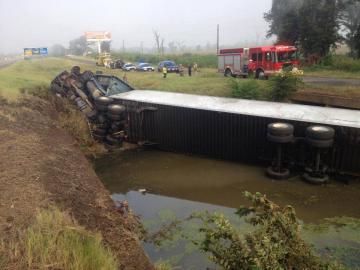 Truck trailer overturned in a ditch beside a rural road, with rescue vehicles nearby.
