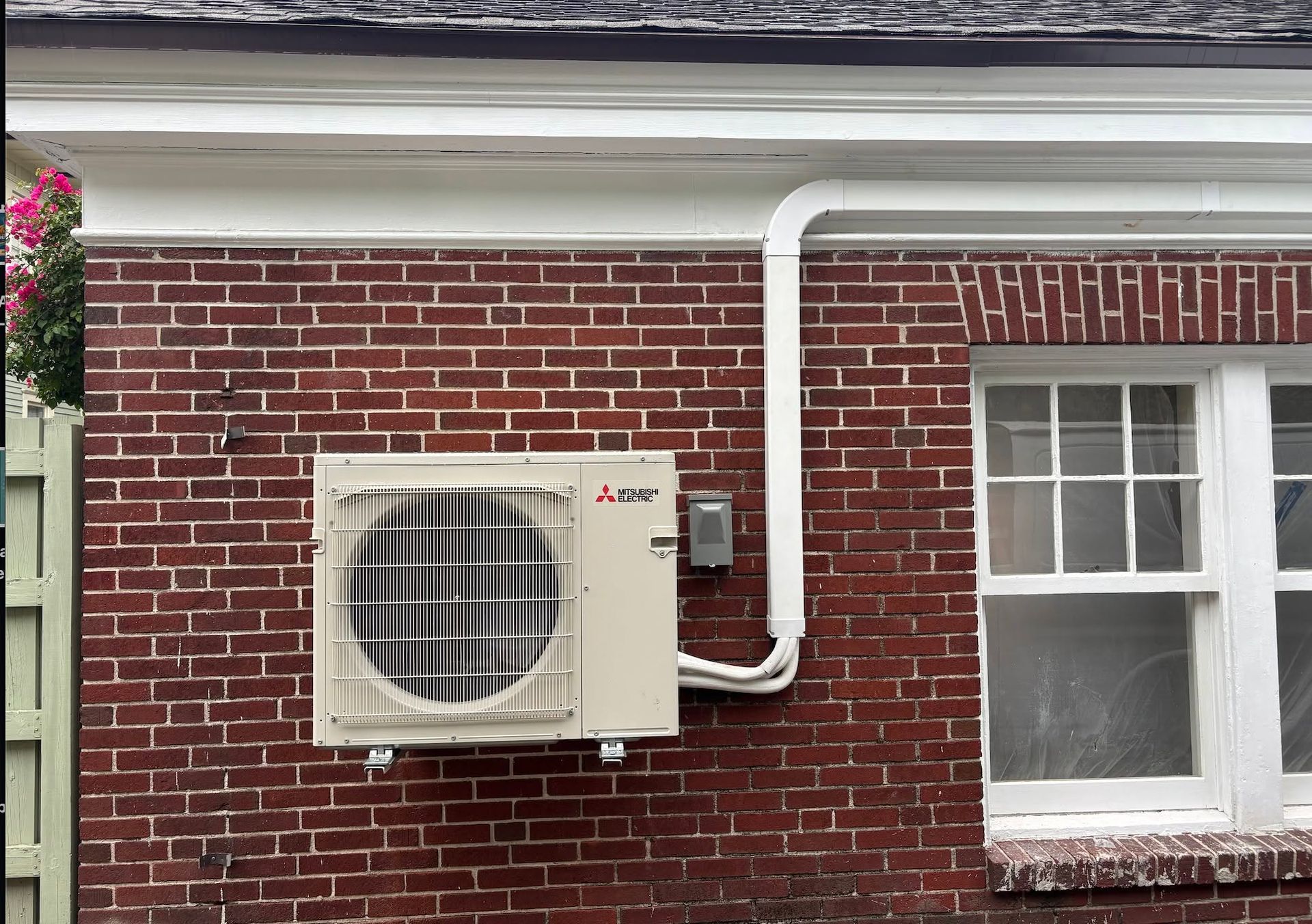 Exterior brick wall with a mounted air conditioner and white piping beside a window