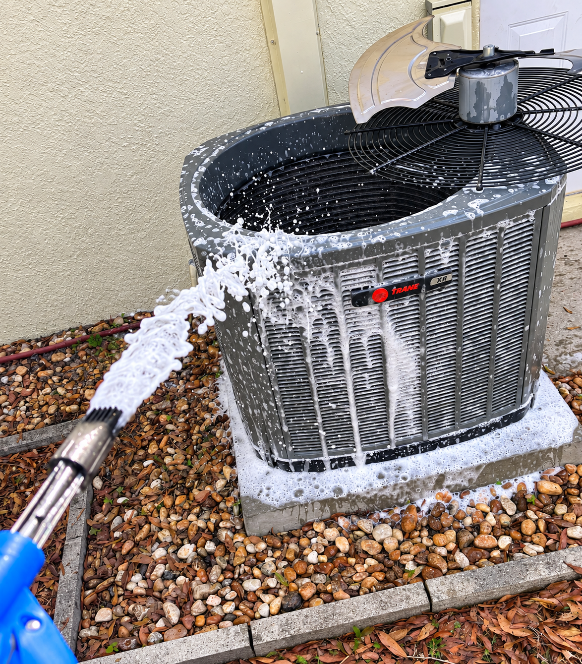 Garden hose spraying water into a dirty outdoor AC unit, rinsing foam off the metal grille.