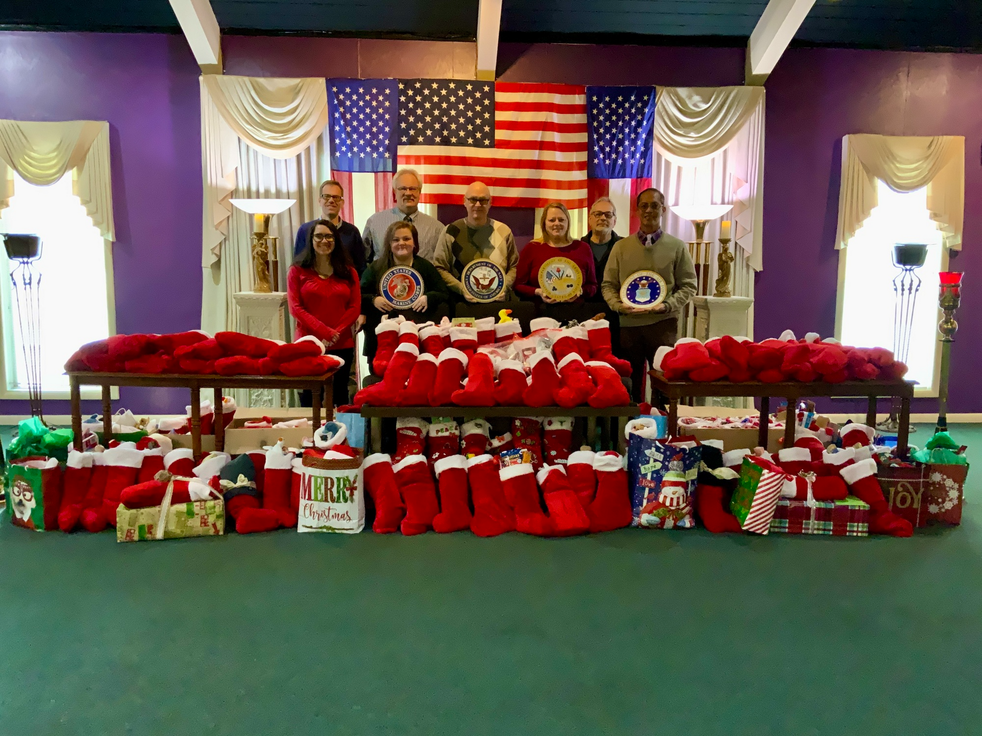 Group of people standing behind piles of Christmas stockings and gifts in front of an American flag and purple backdrop.