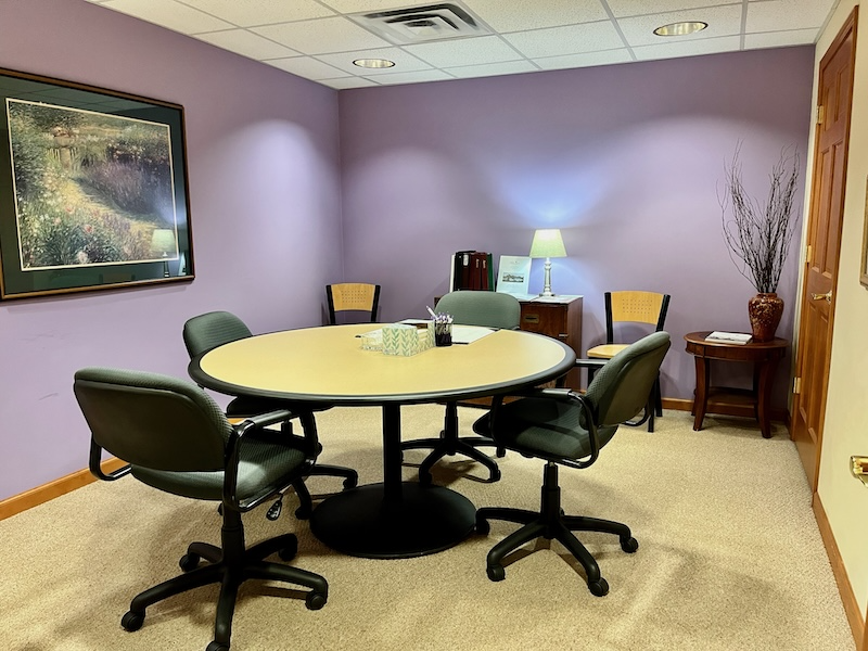 A small office with round table and chairs. Purple walls, beige carpet. Desk with lamp in the back.