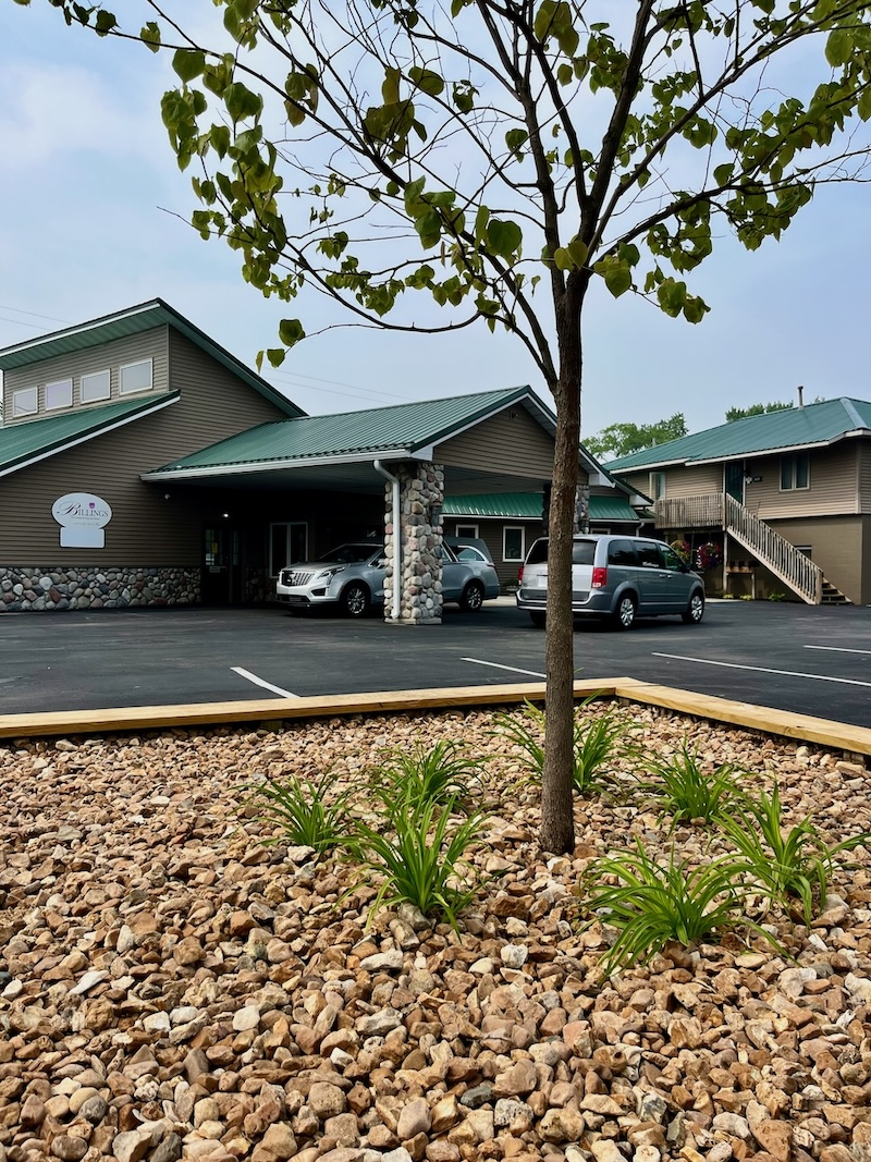 Hotel exterior with cars parked in front, small tree in foreground, and gravel landscaping.