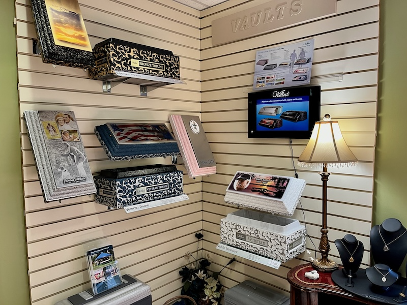 Corner display in a funeral home showcasing vaults, a television, and jewelry. The walls are paneled.