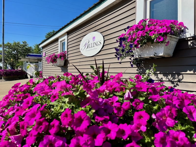 Exterior of a building with bright pink petunias in planters. Building is tan with a logo that says 