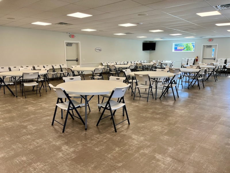 Empty event space with round white tables and folding chairs. Beige carpet, neutral walls, and overhead lighting.