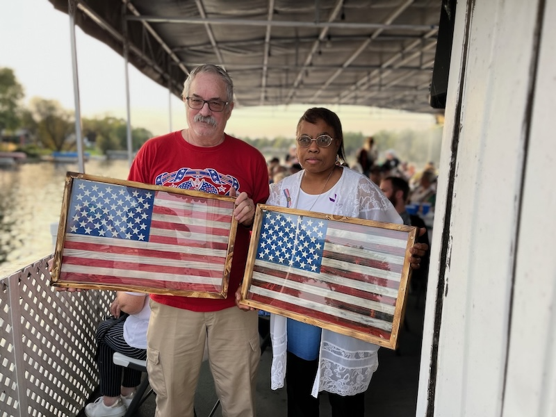 Two people holding framed American flags on a deck, with a body of water in the background.