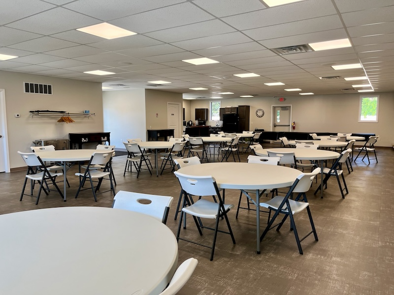 Brightly lit room with round tables and white folding chairs. Likely a community space.