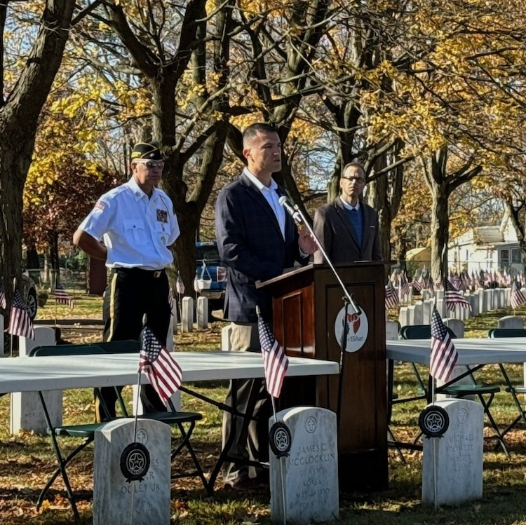 Man speaking at a podium in a cemetery. Two others stand nearby. American flags on graves.