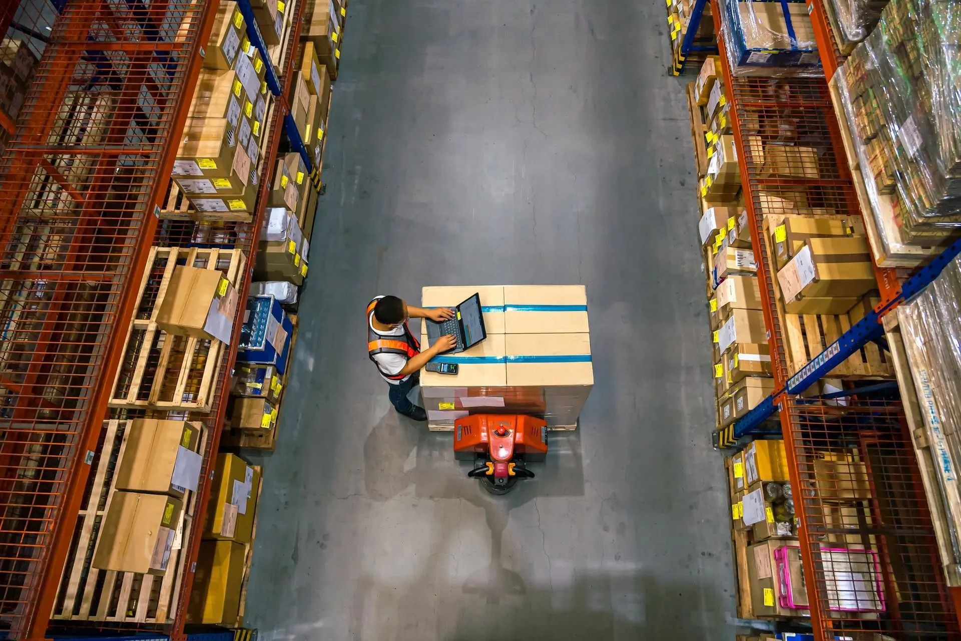 Warehouse worker using laptop on a forklift, surrounded by shelves of boxes.