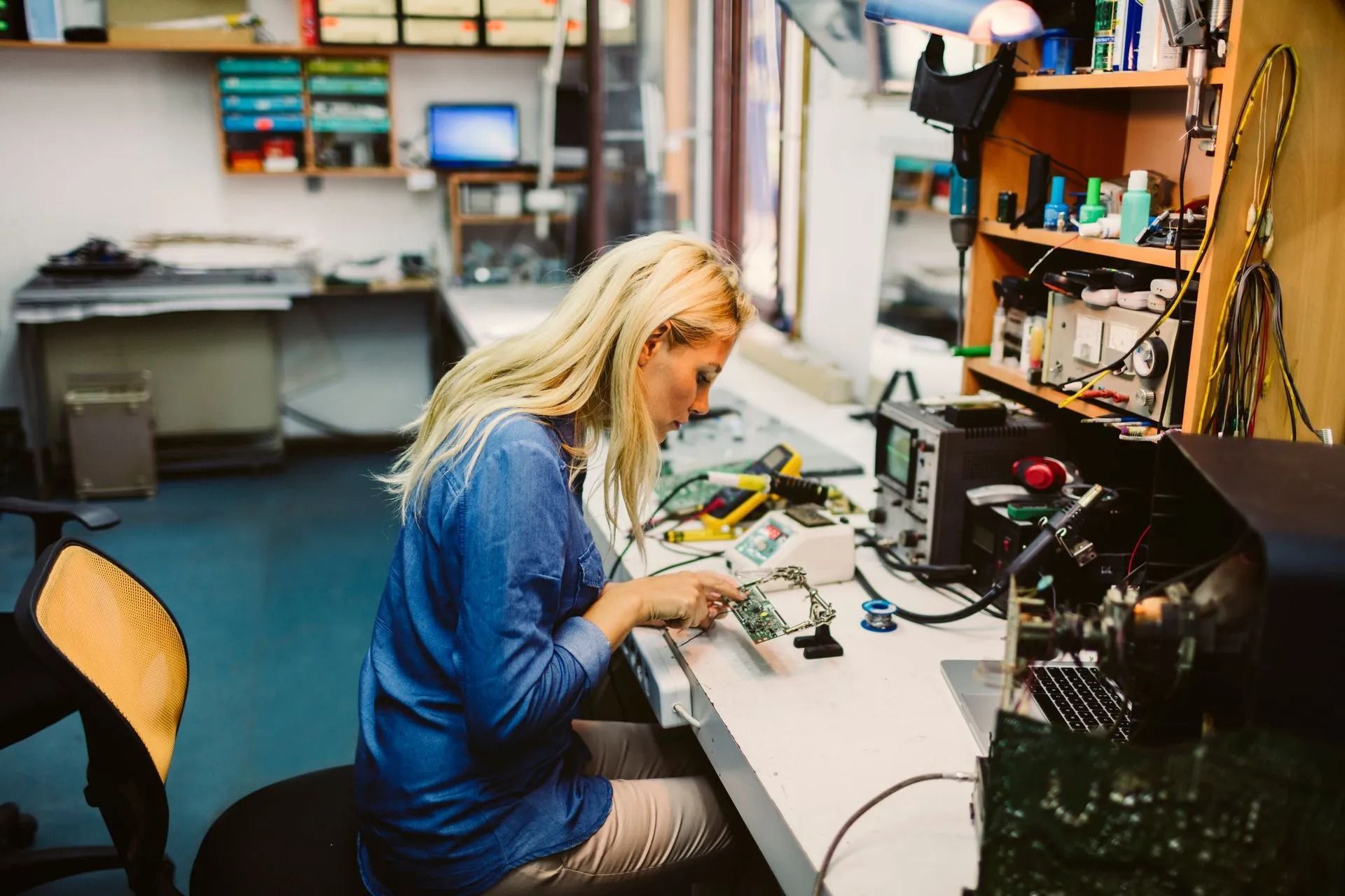 Woman working on circuit board at a workbench, with tools and equipment visible.