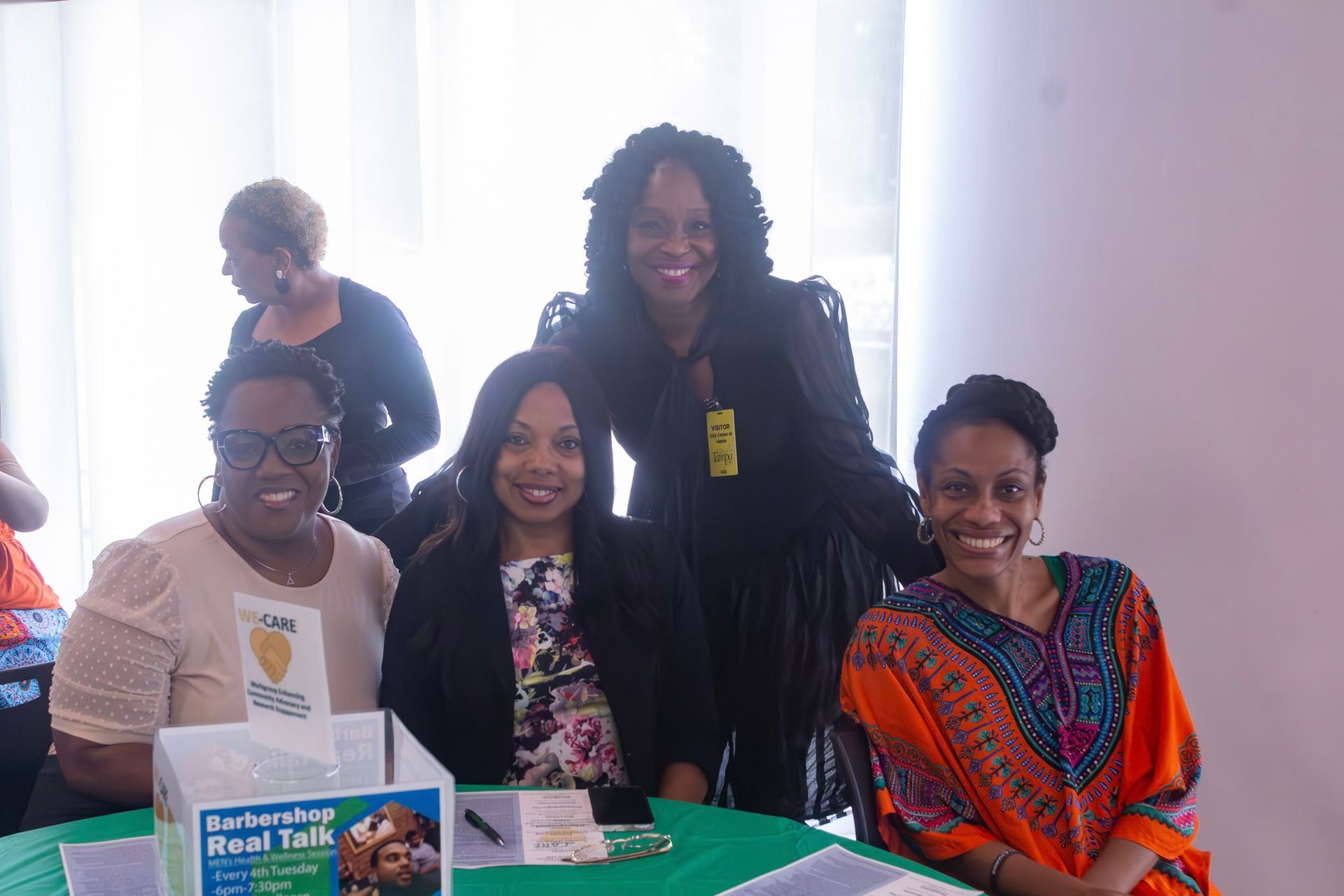 Four women smiling at a table with promotional materials. A fifth woman stands in the background.