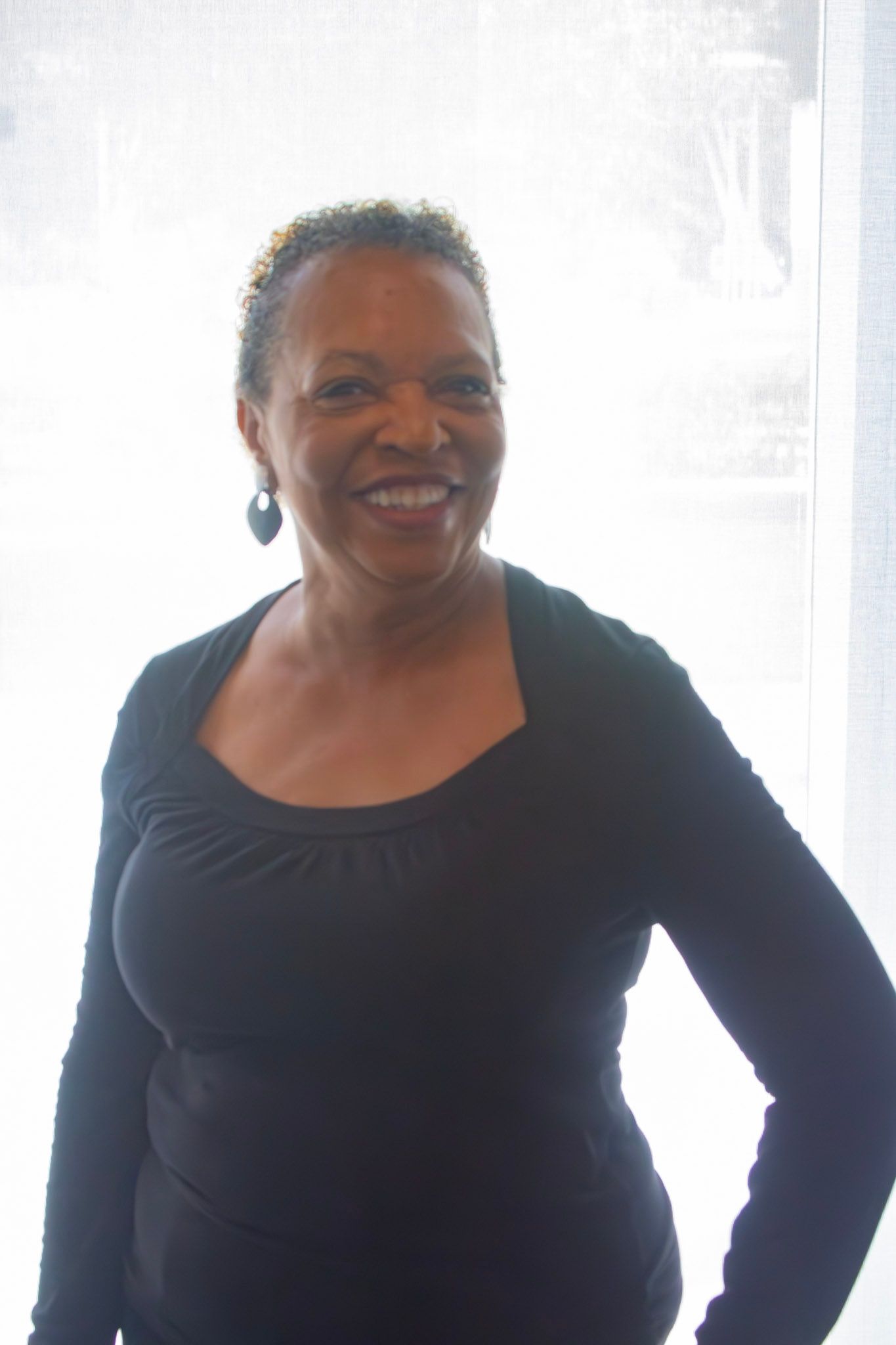 Woman in black top smiling, standing near a window.