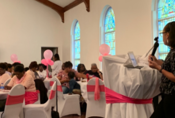 Woman speaking at a pink-themed event in a church. Tables with guests and balloons.