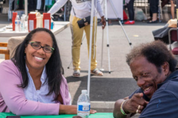 Woman with glasses smiles; man points, laughing. They sit at a table outdoors.
