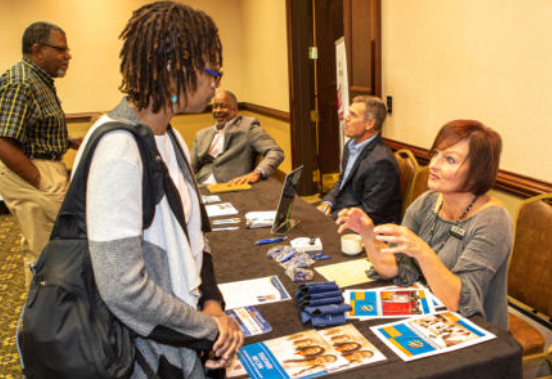 Woman speaking with another at a table, people in background, business conference setting.