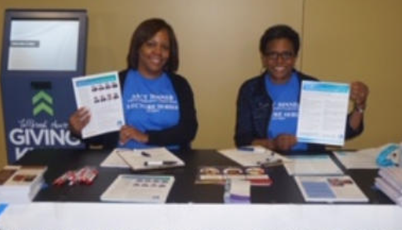 Two people at a table hold papers, smiling. Blue shirts, 