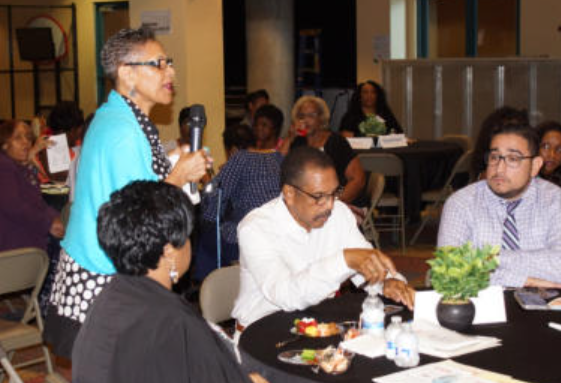Woman speaking into a microphone at a table with other people. Dark setting, some with name tags.