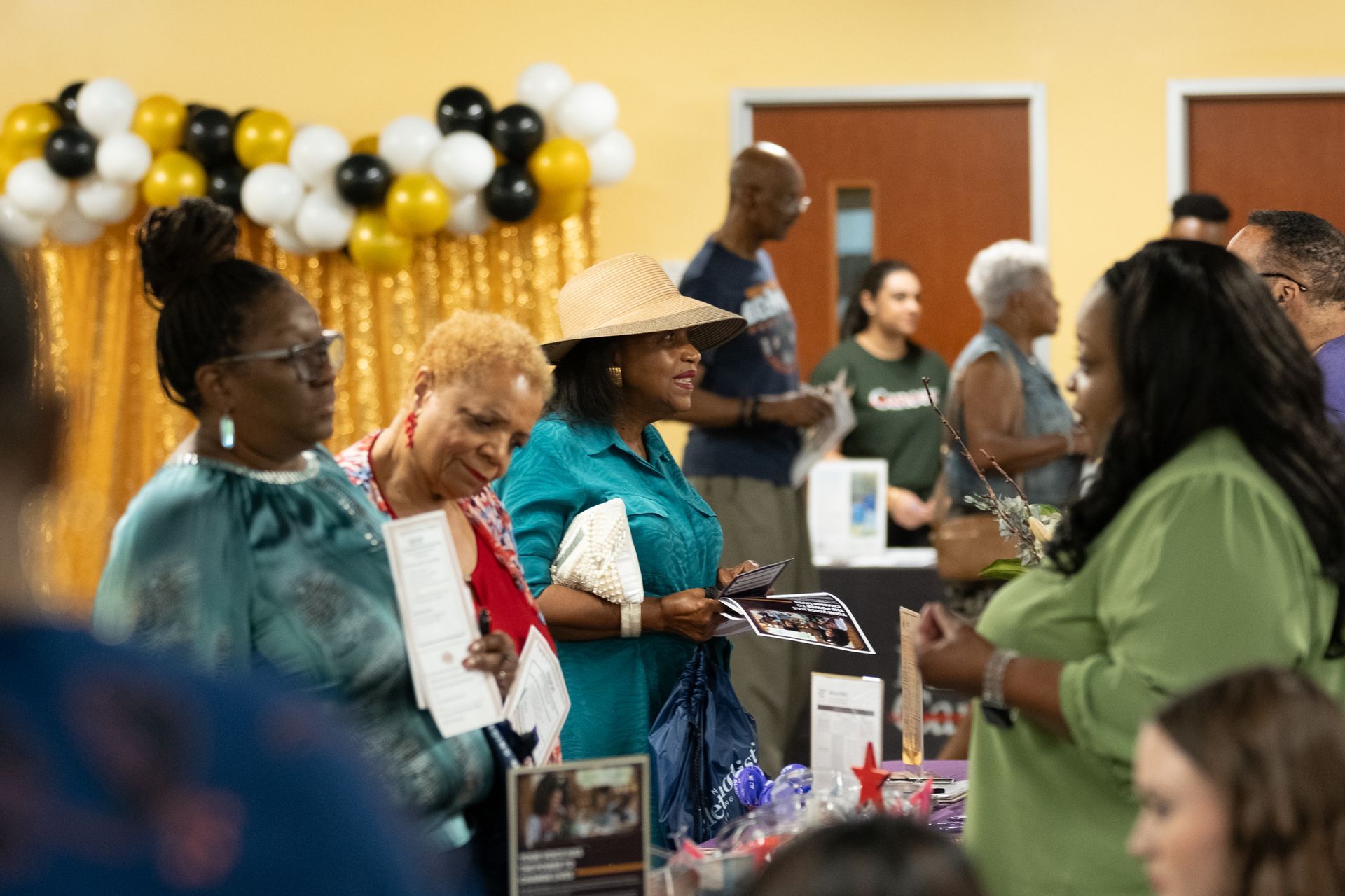 People at an event with balloons and displays. Some are talking and holding items, indoors.