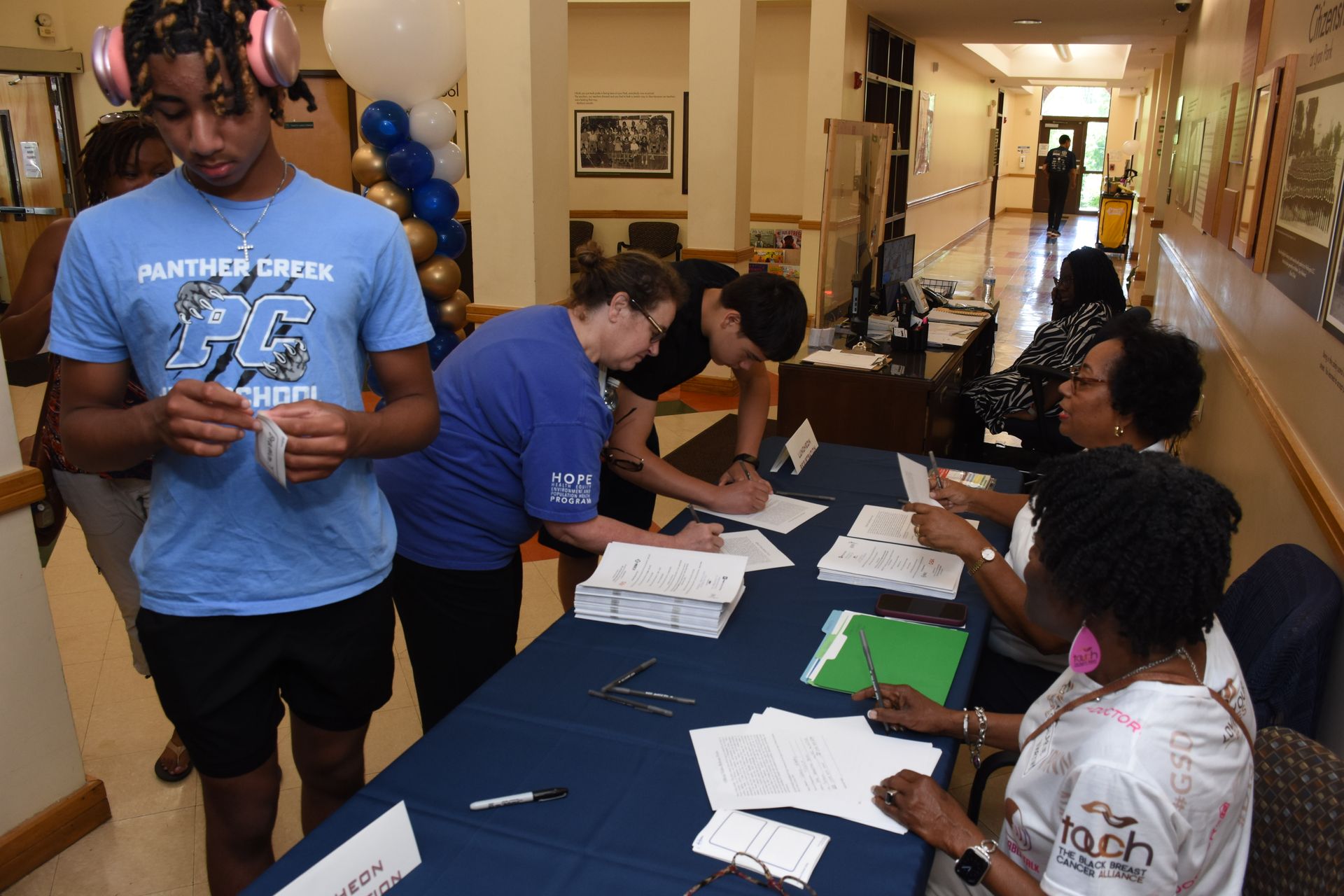 People registering at a table inside a hallway; blue and white balloons, documents, and people.