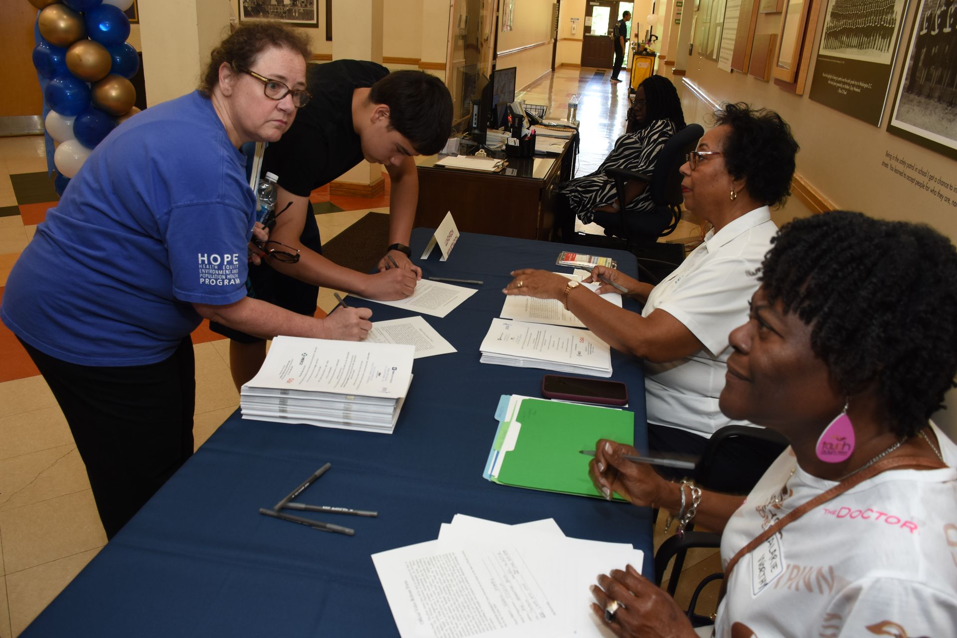 People at a registration table. A woman in blue helps a man sign paperwork. Two women also sign forms.