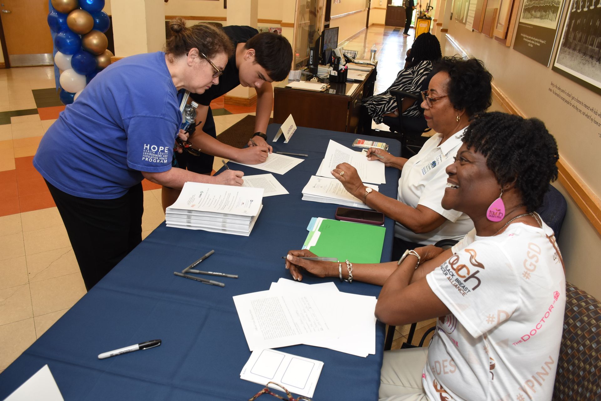 People at a table, signing forms.  Two women and one man helping attendees.  A balloon display is in the background.